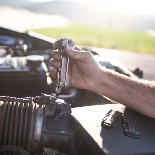 Translation missing: en.A hand using a stainless steel Leatherman Surge® multitool on an air intake, showcasing its practical use in repairs