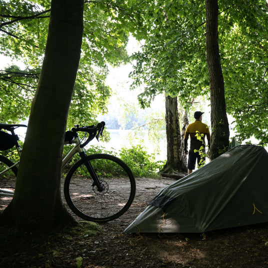 Translation missing: en.A cyclist stands near a tent by the river, surrounded by lush trees in a tranquil camping setting