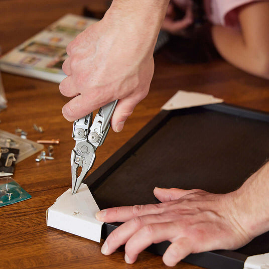 Translation missing: en.A person using a Stainless Steel Leatherman Surge® multitool to fix a picture frame on a wooden table