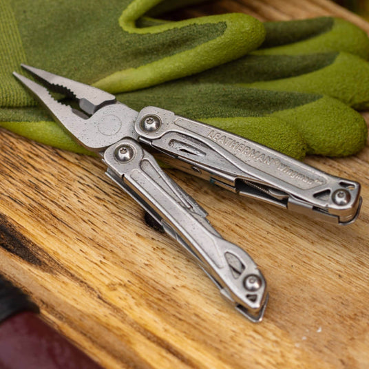 Translation missing: en.Stainless Steel Leatherman Wingman® multi-tool on a wooden surface beside green gloves