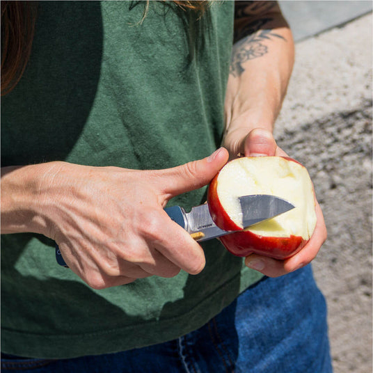 Translation missing: en.Person using a Denim Leatherman Blazer™ knife to slice an apple in a sunny outdoor setting