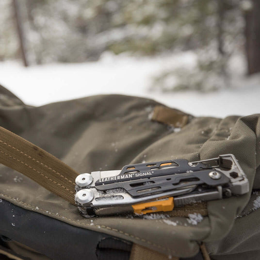 Translation missing: en.Gray Leatherman Signal® multi-tool resting on a backpack strap in a snowy environment