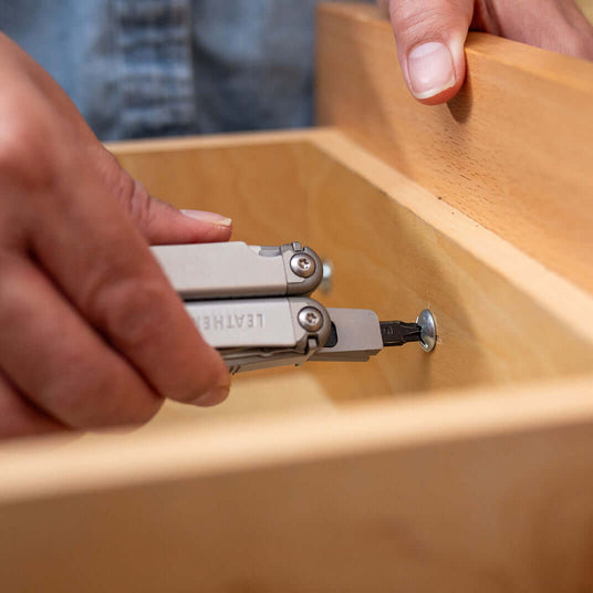 Translation missing: en.Hand using a Stainless Steel Leatherman Wave® Plus multitool on a wooden drawer, showcasing its practical features