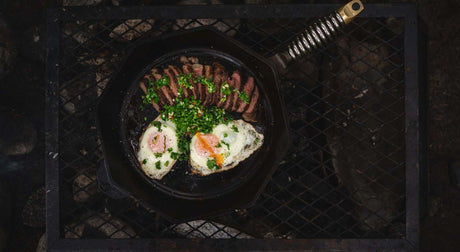 Translation missing: en.A cast iron skillet on a grill, featuring sunny-side up eggs, sliced meat, and fresh herbs