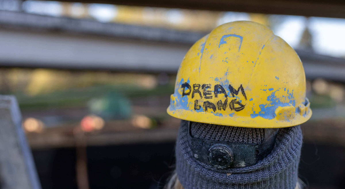 Close-up of a yellow hard hat with 'Dream Land' painted on it, set against a blurred construction background.