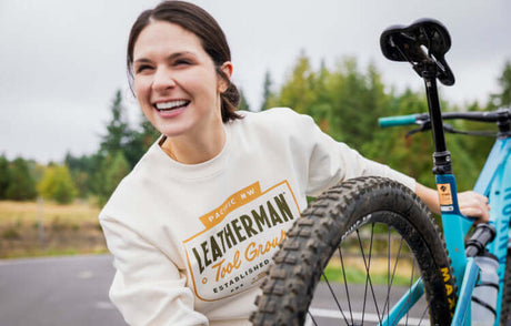 Translation missing: en. Woman wearing a Natural Raw Leatherman Pacific Northwest Crewneck sweatshirt while holding a mountain bike