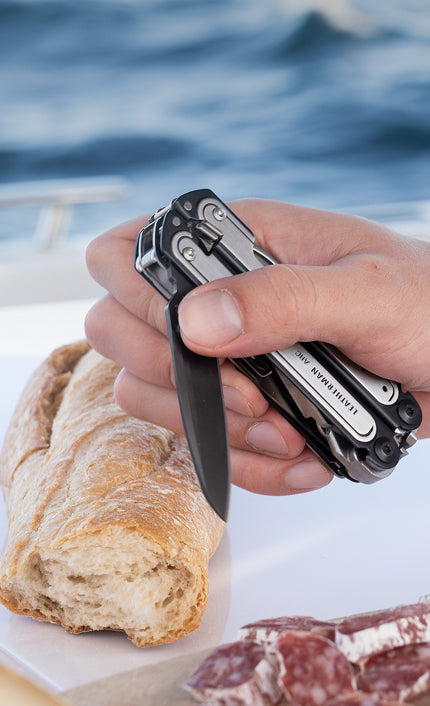 Translation missing: en.Hand holding a stainless steel Leatherman Arc multitool above a loaf of bread and cured meats on a boat