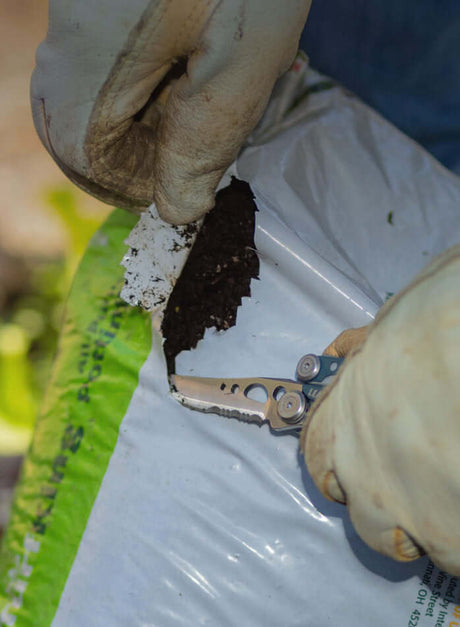 Translation missing: en.Person using a Denim Skeletool® to open a bag of soil, showcasing the utility of this versatile tool in gardening tasks