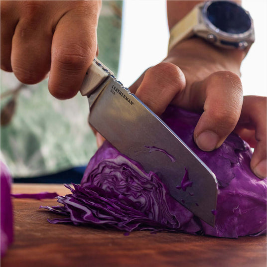 Translation missing: en.Person using an Alpine Leatherman Rustle™ knife to chop purple cabbage on a wooden cutting board