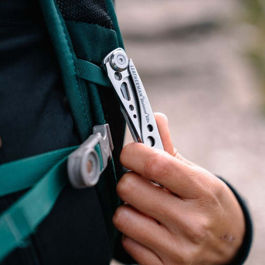 Translation missing: en.Hand holding a Stainless Steel Leatherman Skeletool® KBX multi-tool attached to a backpack strap outdoors