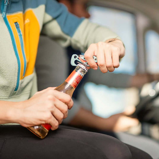 Translation missing: en.Person using a Paradise Skeletool® CX to open a bottle in a car, showcasing its multi-tool functionality