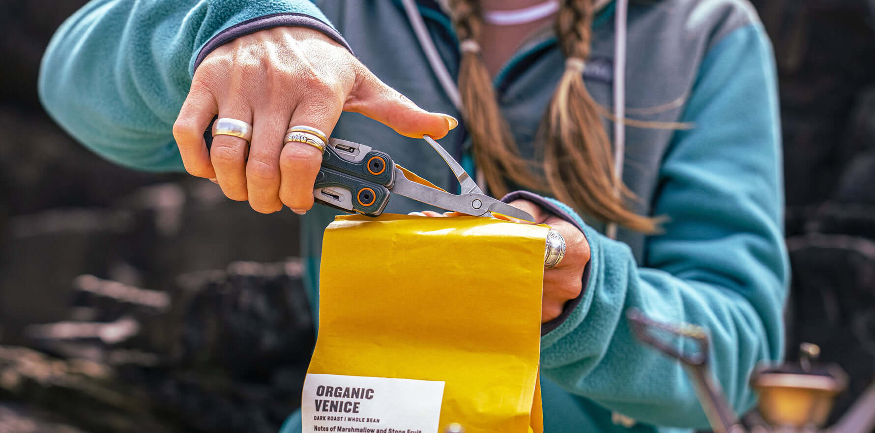 Person using the scissors on a Cascadia Leatherman Wave® Alpha to cut open a yellow bag of coffee beans outdoors.