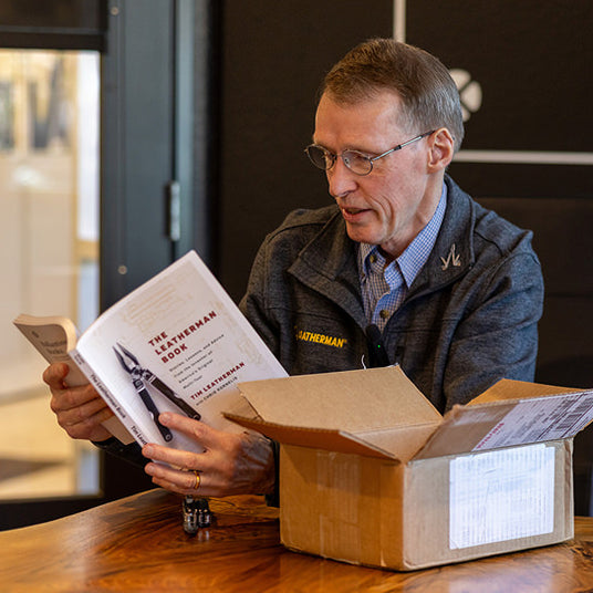 Translation missing: en.Tim Leatherman sits at a table reading The Leatherman Book, with an open cardboard box and a multitool beside him