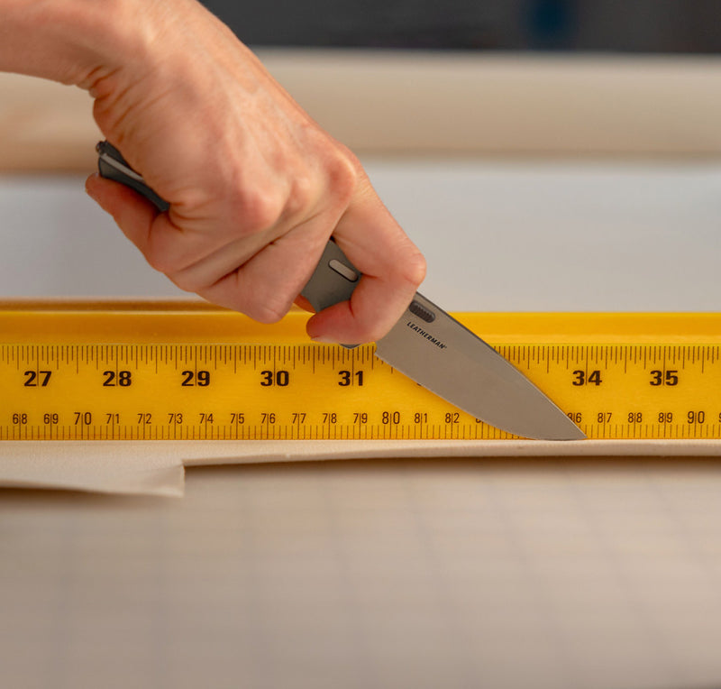 Translation missing: en.Close-up image of a hand using a Leatherman folding knife to cut fabric along a yellow measuring ruler on a gridded cutting mat, illustrating precision and utility in a workspace or crafting environment