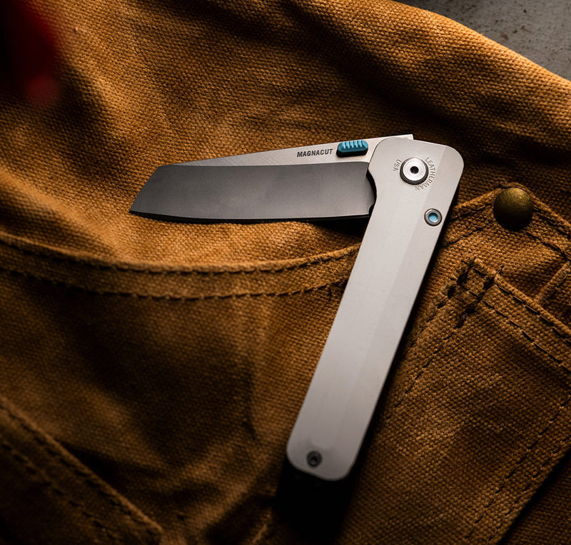 Translation missing: en.Close-up image of a Leatherman folding knife with a straight-edge blade engraved with “MAGNACUT,” resting on the front pocket of brown canvas workwear. The knife features a sleek, silver handle with a blue locking mechanism and accent, showcasing a rugged yet refined design