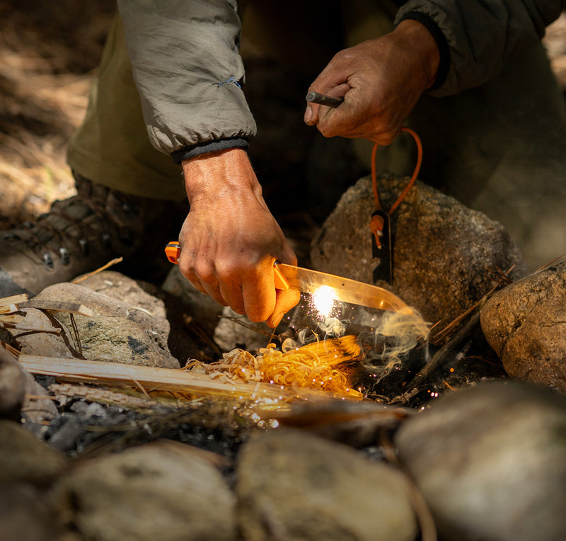 Translation missing: en.Person igniting a fire using a ferro rod and a Leatherman fixed-blade knife with an orange handle, creating sparks over kindling in a rocky outdoor campsite setting