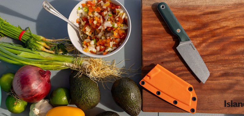 Translation missing: en.Overhead lifestyle image showcasing a Leatherman Rustle Evergreen knife on a wooden cutting board, with its bright orange sheath nearby. Surrounding the board are fresh vegetables including avocados, scallions, red onion, limes, and a bowl of freshly chopped salsa, emphasizing an outdoor food prep scene