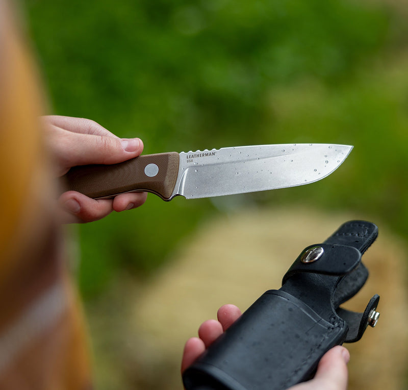 Translation missing: en.A person holding a Leatherman fixed-blade knife with a brown handle and a wet blade, positioned above a black leather sheath in the other hand. The background features soft-focus greenery, suggesting an outdoor or wilderness setting