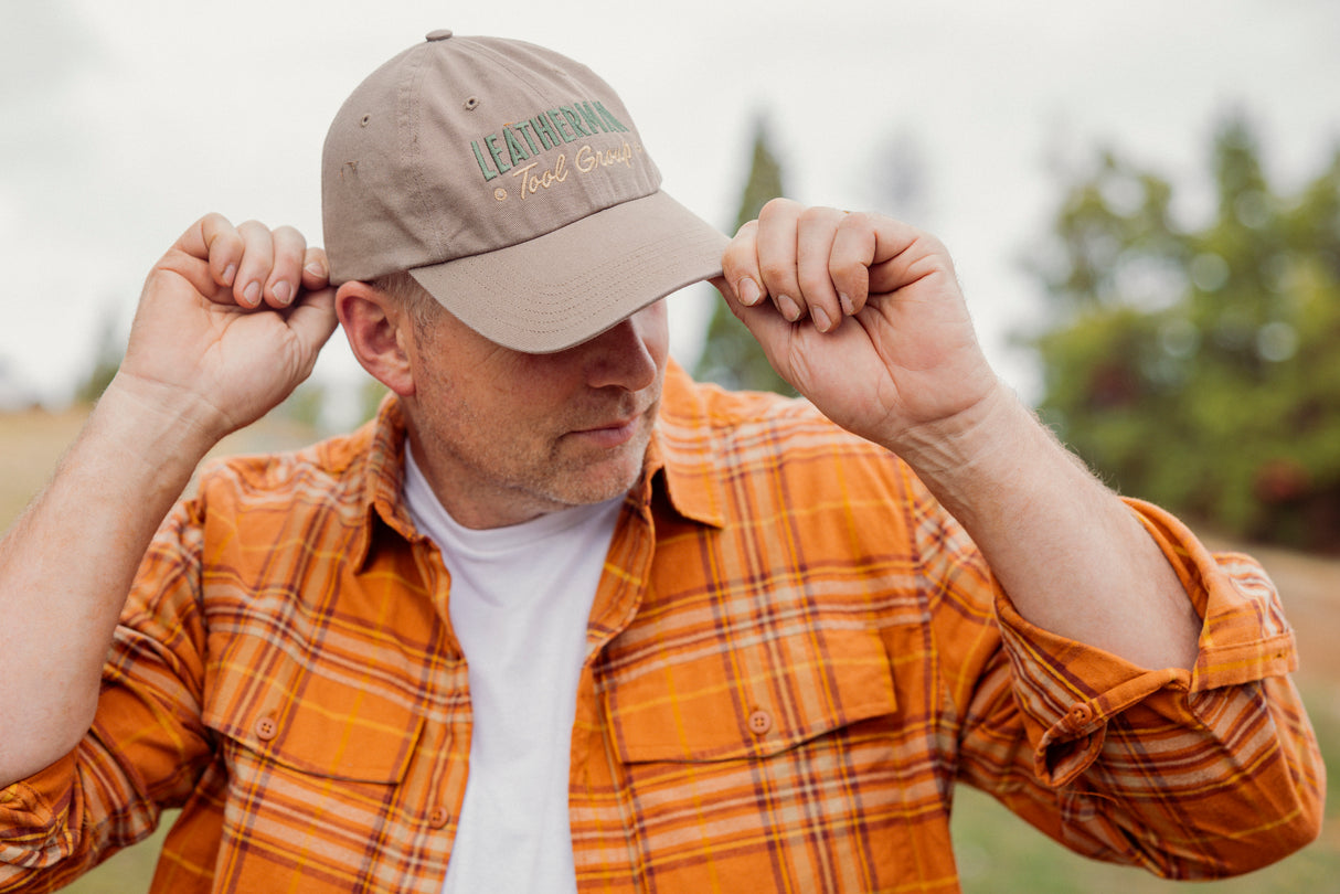 Translation missing: en.A person wearing a Leatherman Dad Hat in Driftwood color adjusting the brim while outdoors, dressed in an orange plaid shirt