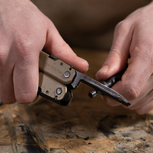 Translation missing: en.Close-up of hands using a Coyote Tan Leatherman Super Tool® 300M multi-tool on a workbench