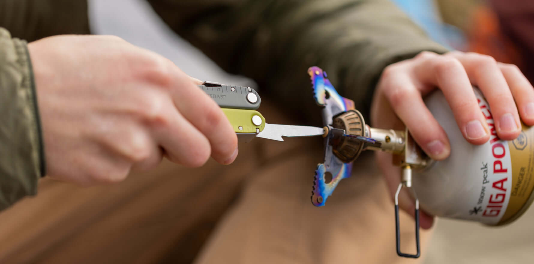 A person using a Mossy Slate Leatherman Rebar® multi-tool to adjust a camping stove, showcasing its functionality outdoors.
