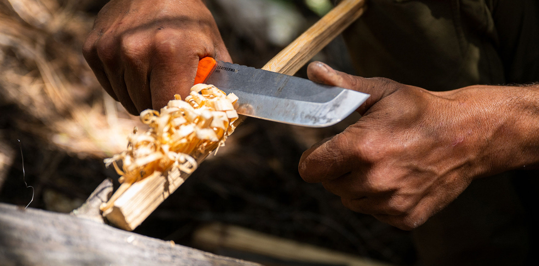 Close-up of hands using an Orange Pop Leatherman Pioneer™ knife to carve wood shavings outdoors.