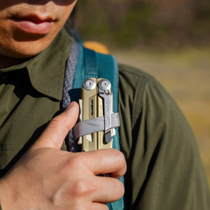 Translation missing: en.Person securing a Signal Sandstorm multitool to a backpack shoulder strap with a gray loop outdoors