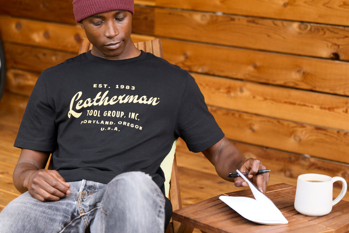 Translation missing: en.A person wearing a black Leatherman Vintage Tee sitting on a wooden deck chair, opening a notebook beside a mug of coffee